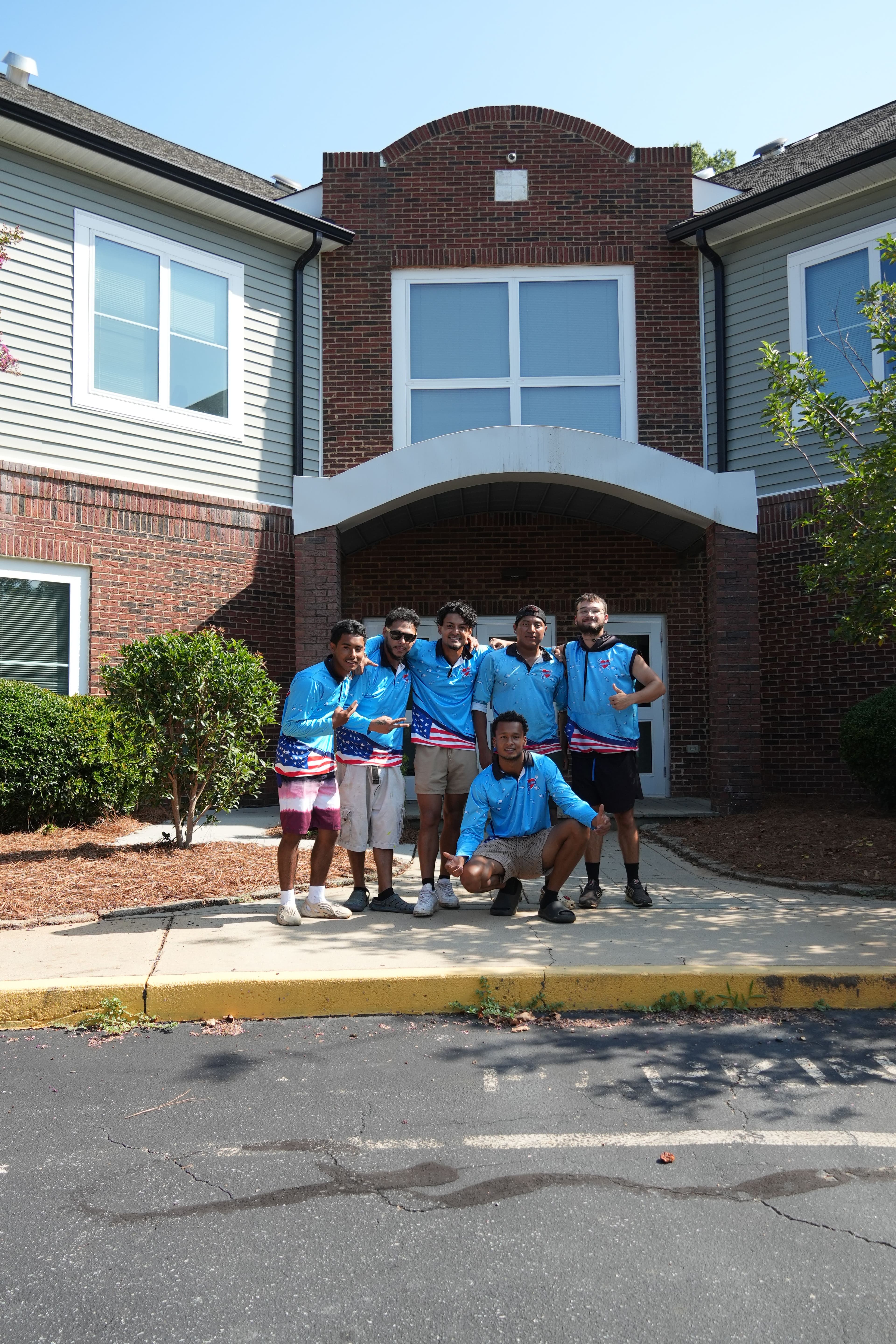 Group of seven friends in matching blue shirts posing in front of a brown brick building.