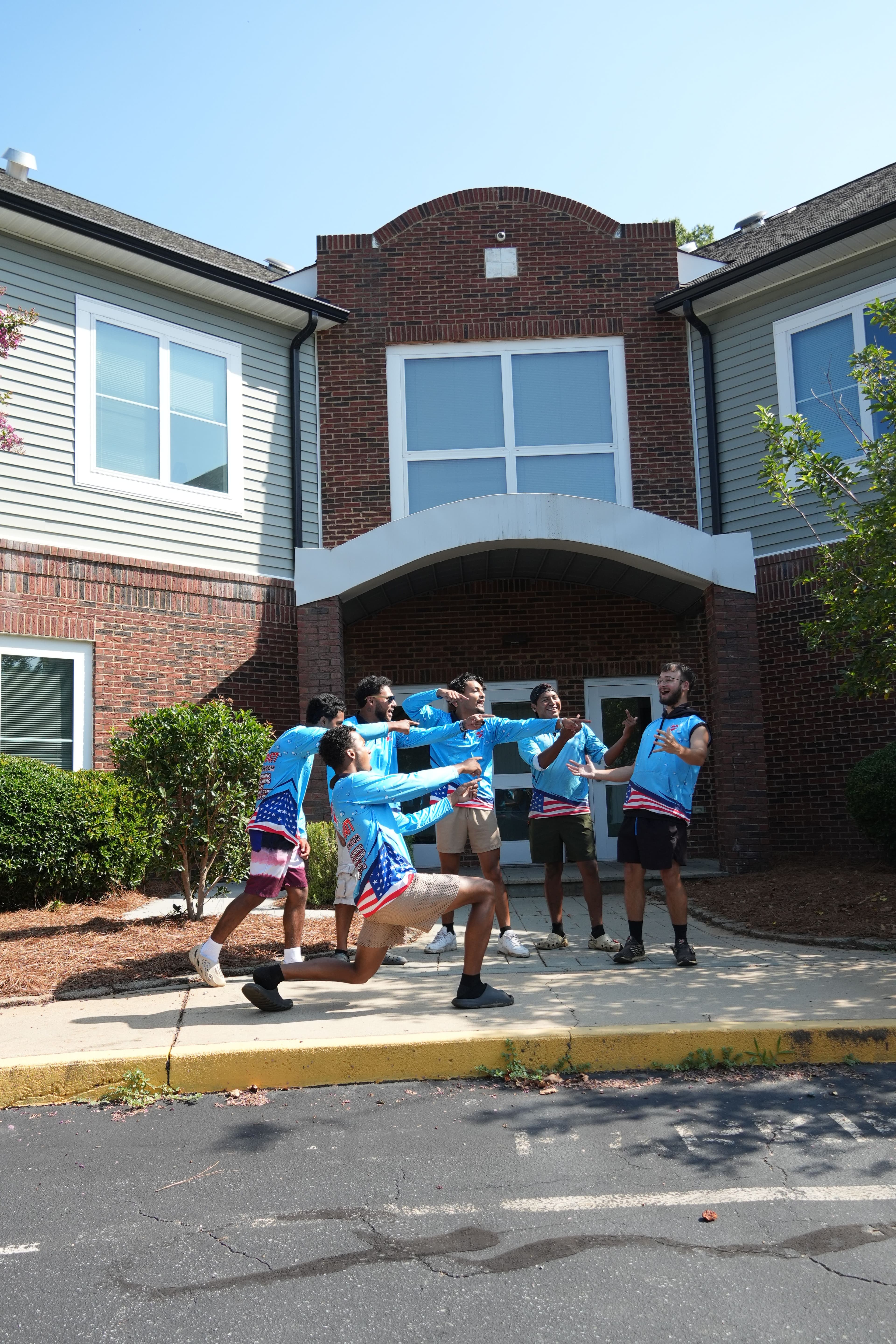 Group of six young people in blue shirts posing outside a residential building on a sunny day.