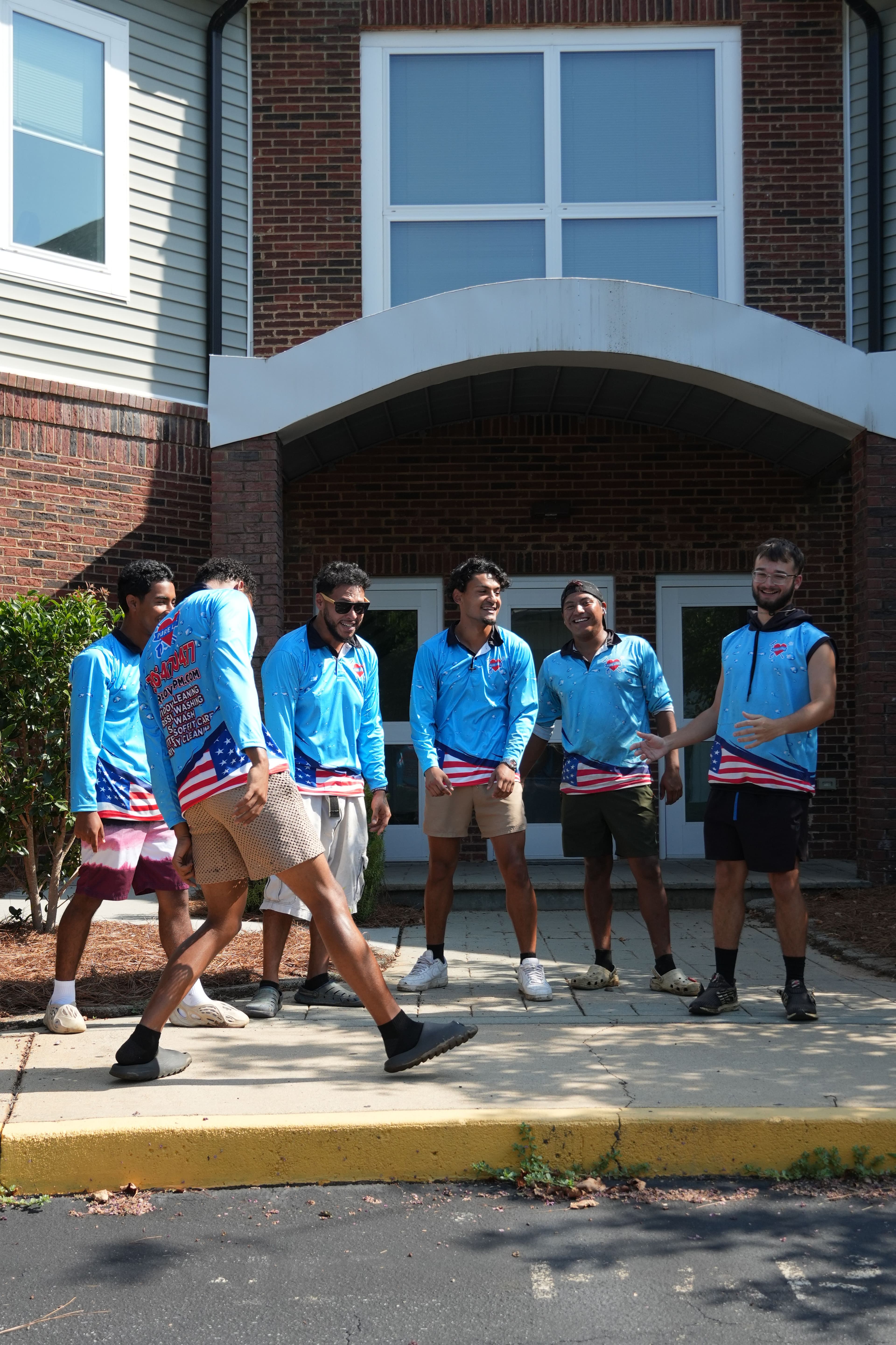 Group of six men in blue shirts with American flag designs, posing outside a brick building.